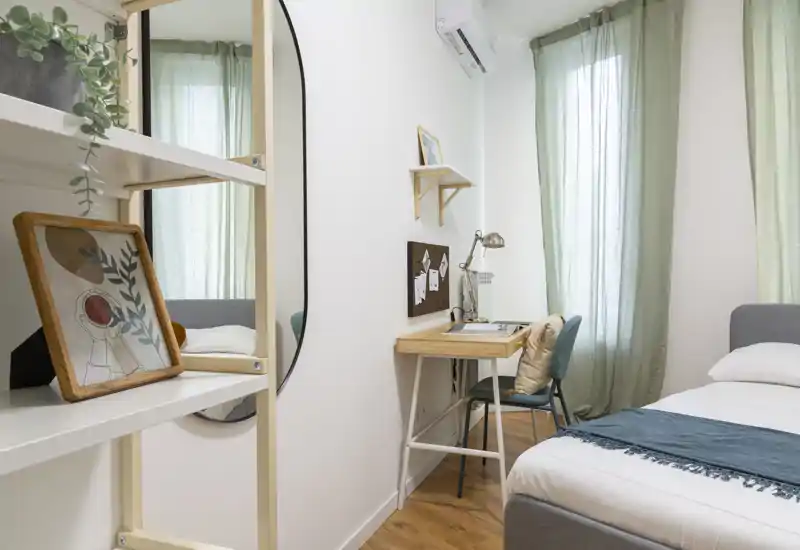 Camera da letto luminosa e compatta con letto ben fatto, una piccola scrivania in legno e tende verdi che filtrano la luce naturale — perfetta per un soggiorno accogliente in città.