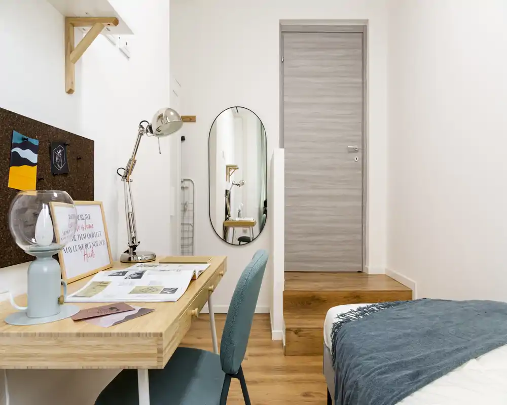 Bright, modern bedroom corner with a neatly made bed, a wooden desk and teal chair — ideal for work and rest.