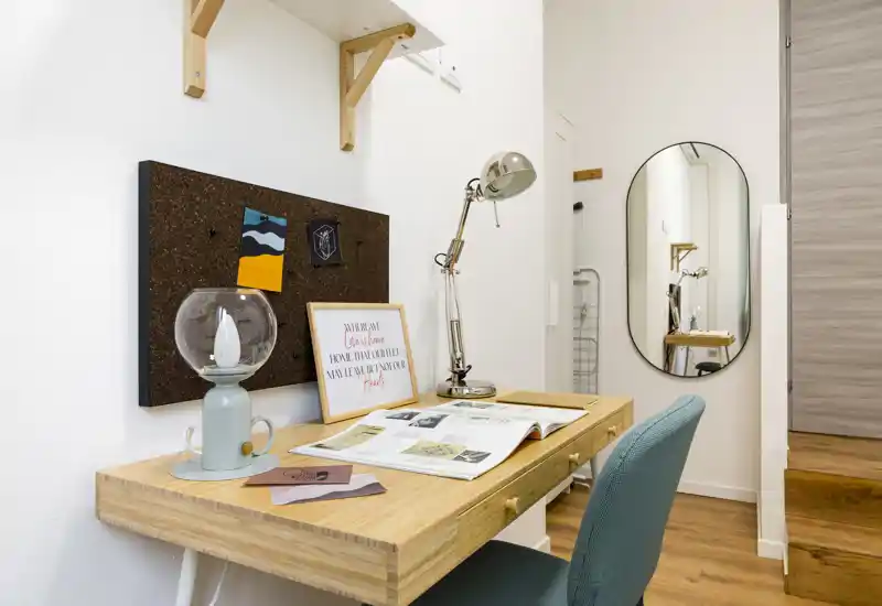 Rincón de trabajo en la habitación con escritorio de madera, lámpara de lectura, tablero de corcho y espejo; suelo cálido y silla cómoda.
