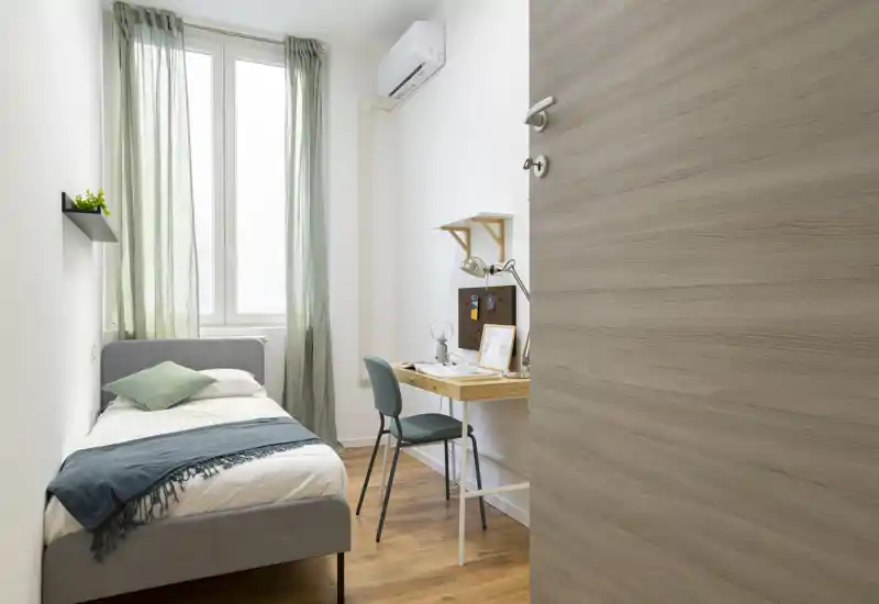 Habitación individual ordenada con cama gris acogedora, toques en verde y un pequeño escritorio de madera junto a una ventana luminosa — ideal para estudiante o persona sola.