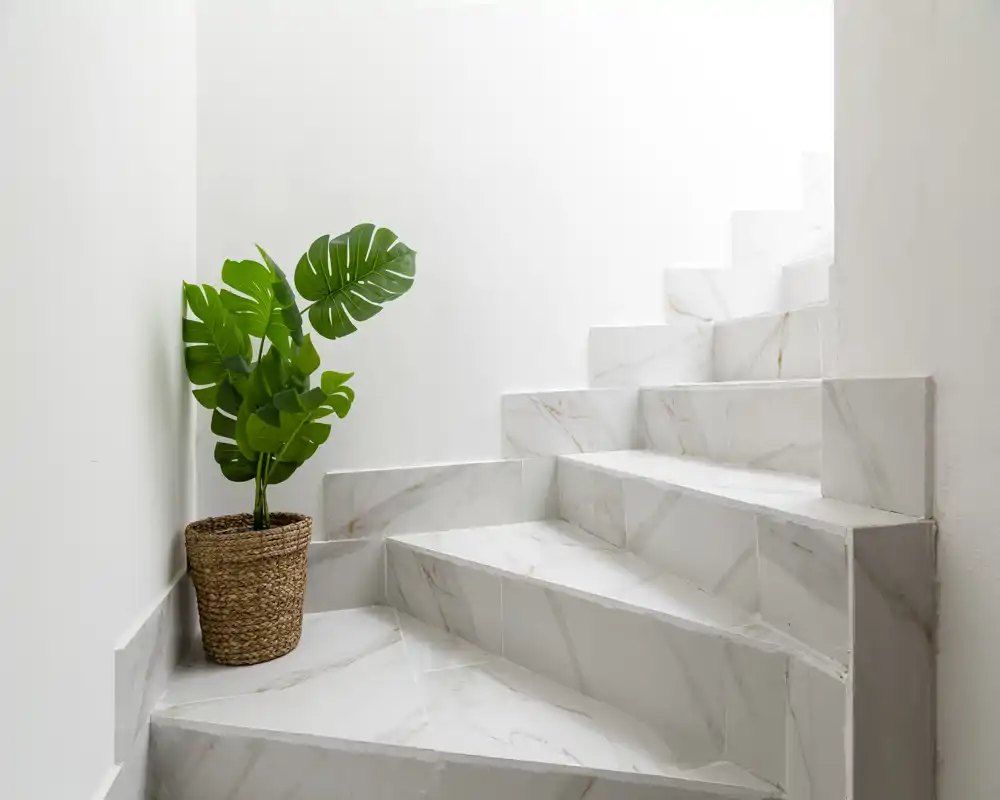 Scala minimale in marmo che sale accanto a una parete bianca, decorata con un vaso intrecciato e fogliame verde per un ingresso o vano scale moderno e pulito.