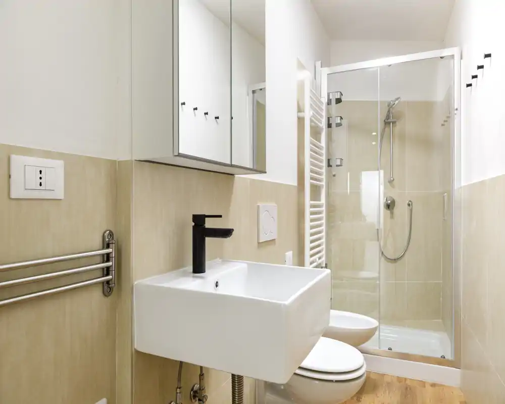 Bagno moderno e luminoso con lavabo a parete, wc e doccia con box in vetro. Piastrelle neutre e arredi essenziali creano un aspetto pulito e contemporaneo.