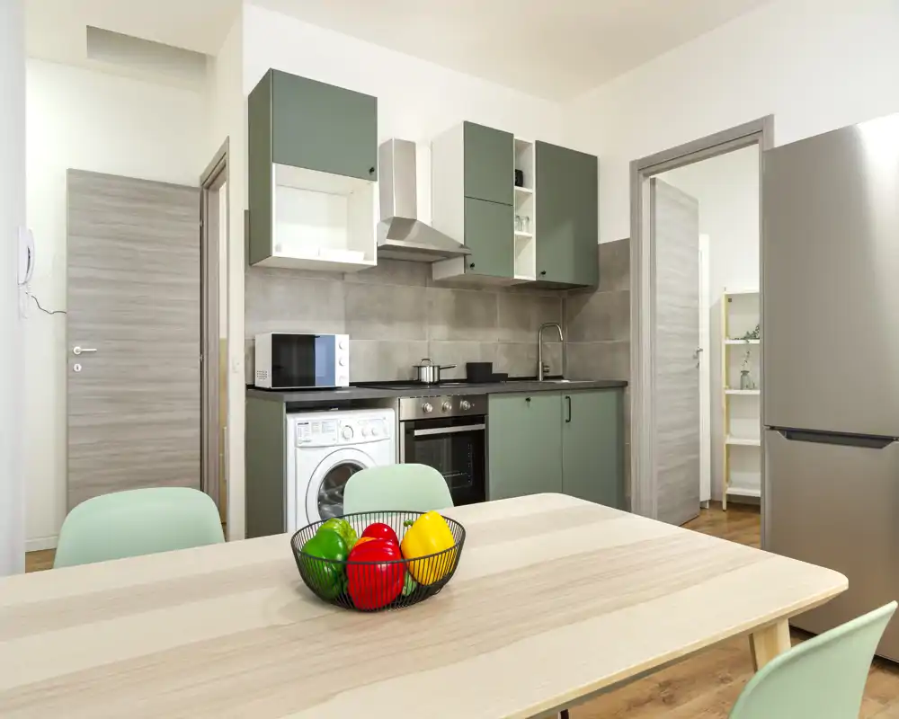Cucina moderna e compatta con mobili verdi, forno e lavatrice integrati, tavolo in legno e sedie pastello — luminosa, ordinata e funzionale per soggiorni brevi.