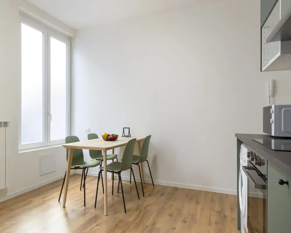 Zona pranzo moderna e luminosa con tavolo in legno semplice, quattro sedie verdi e una grande finestra che illumina l'ambiente — perfetta per pasti informali.