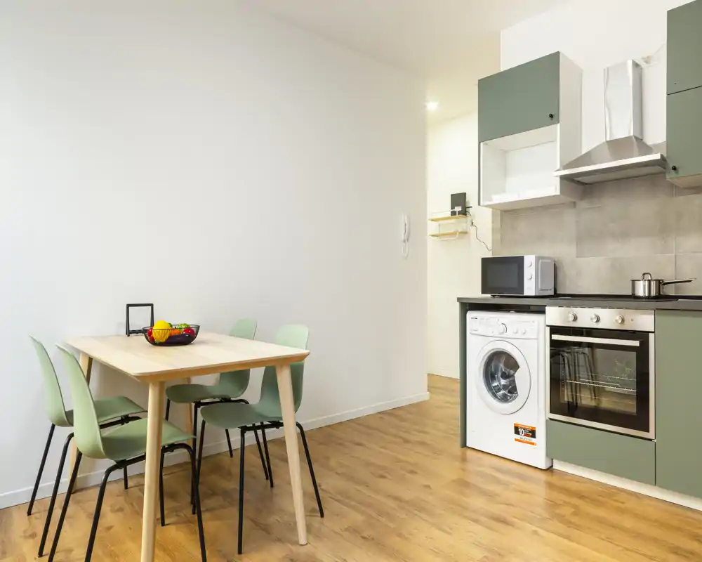 Cucina compatta e luminosa con mobili verde oliva, forno integrato, lavatrice e tavolo da pranzo con quattro sedie — perfetta per un monolocale moderno.