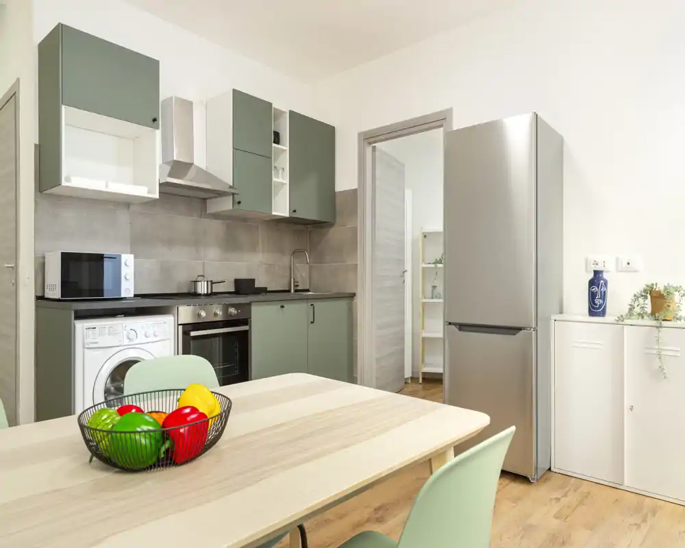 Cucina moderna e luminosa con ante verdi, frigorifero inox e tavolo in legno con frutta colorata — ambiente pulito e funzionale per cucinare e pranzi informali.