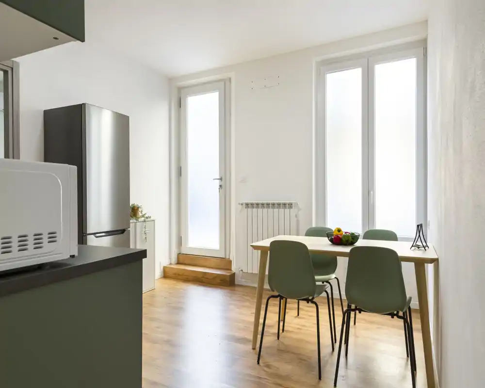 Angolo cucina moderno e luminoso con un piccolo tavolo da pranzo e quattro sedie verdi, frigorifero in acciaio e luce naturale da ampie finestre e porta.