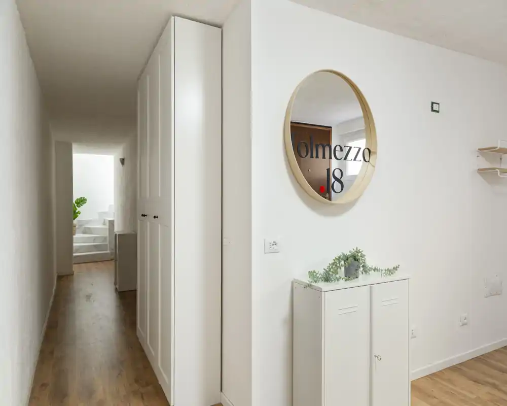Ingresso luminoso e minimal con pavimento in legno, un mobiletto bianco e uno specchio tondo che riflette l'area della porta d'ingresso.