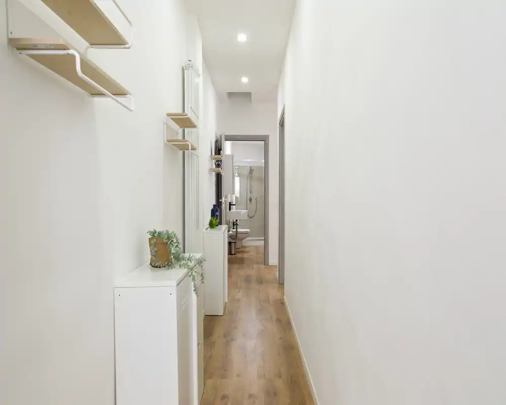 Corridoio stretto e luminoso con pavimento effetto legno, pareti bianche e piccoli mobili contenitori — ingresso ordinato che conduce al bagno.
