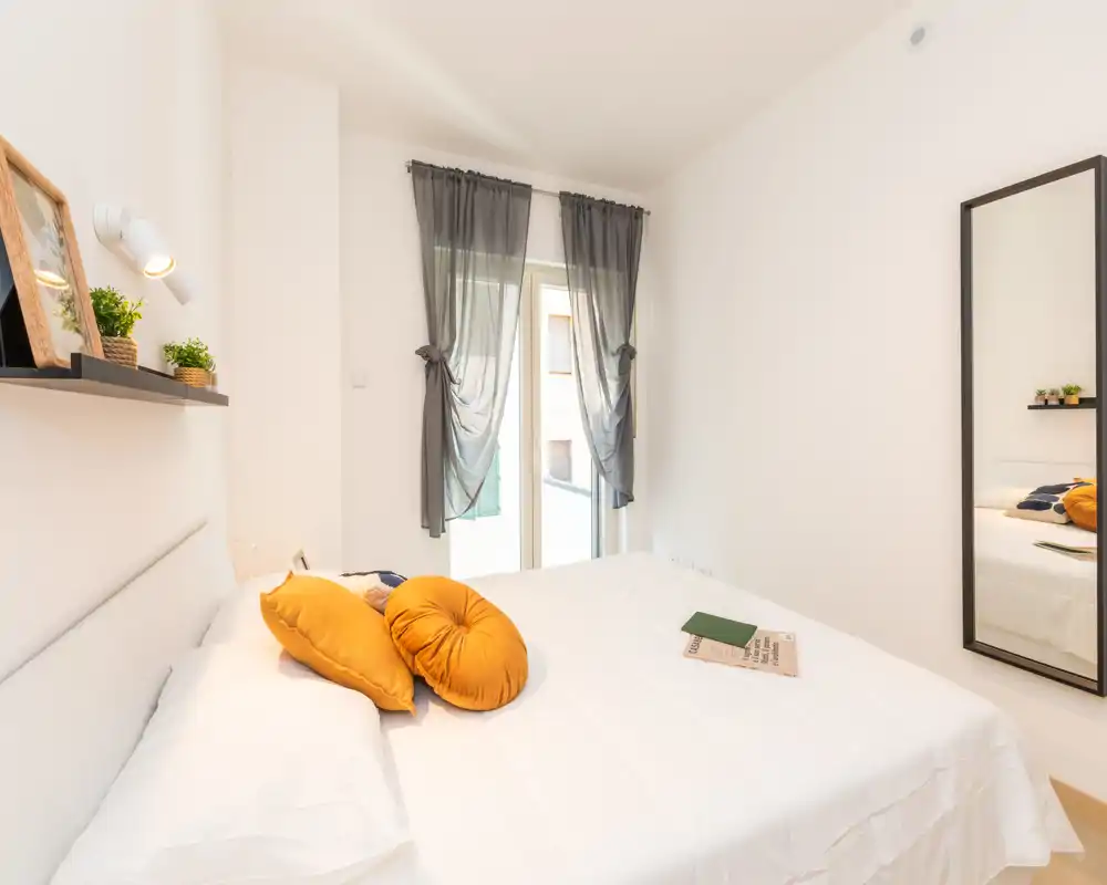 Bright, minimalist bedroom with a large white bed, warm ochre accent cushions, a full-length mirror and a balcony window dressed with sheer gray curtains — well staged and welcoming for rental photos.