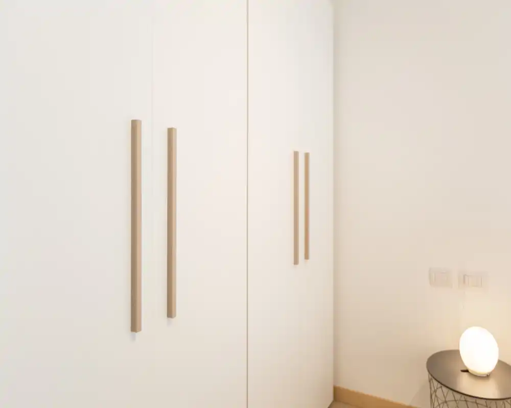 Minimal bedroom corner featuring a tall white wardrobe with long wooden handles and a small round bedside table with a glowing lamp; neutral tones and soft lighting create a calm atmosphere.