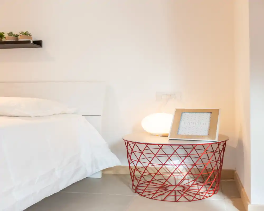 Bright, minimal bedroom corner with a white bed, soft linens and a modern wire-frame bedside table topped by a warm lamp and framed print.