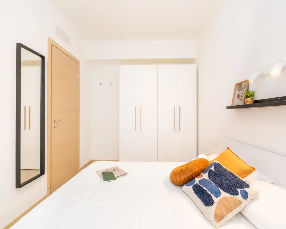 Bright, modern bedroom with a large white bed, decorative cushions and a wall shelf with a lamp and plant. Clean minimal styling and light wood accents create a calm, welcoming space.