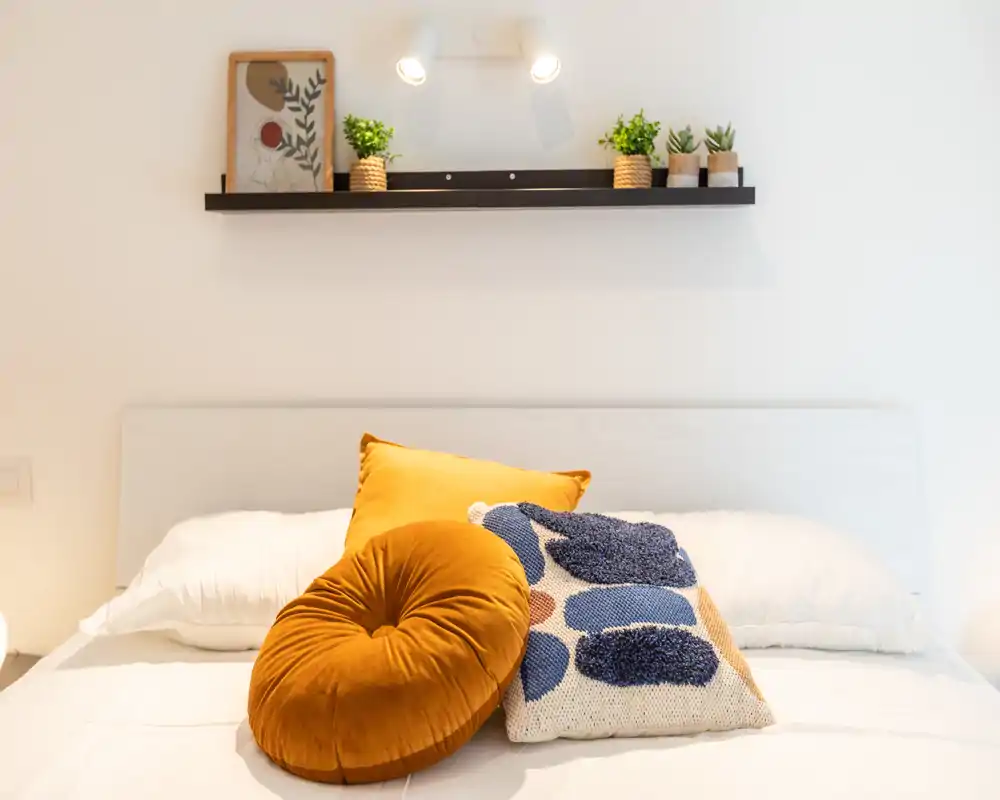 Bright, styled bedroom with a white headboard, layered pillows and decorative cushions in warm ochre and textured blue tones; a small shelf with plants and art above adds a cozy touch.