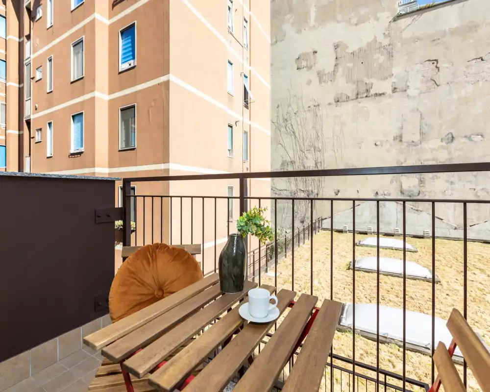Sunny private balcony with a small wooden table and two chairs, perfect for morning coffee. Views overlook an inner courtyard and neighboring building façades.