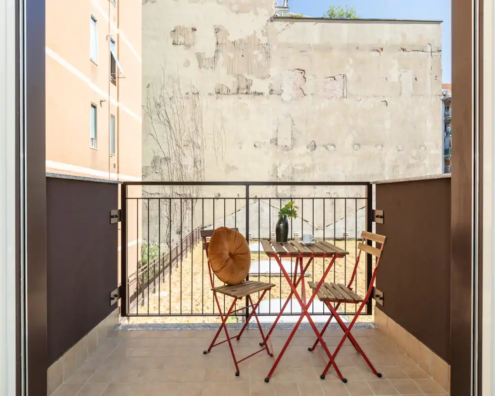 Sunny private balcony with a small wooden bistro table and two chairs, tiled floor and railing overlooking a quiet courtyard — ideal for morning coffee.