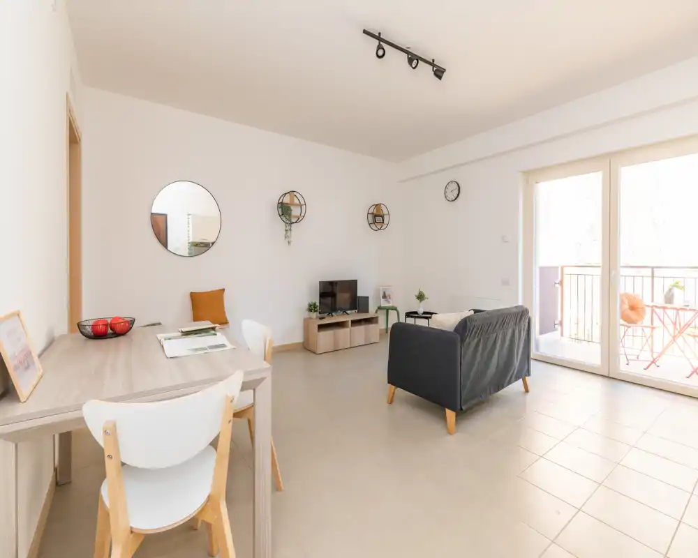 Bright, modern living room with a compact sofa, TV unit, dining table and access to a sunny balcony—clean minimal styling and natural light make it inviting.
