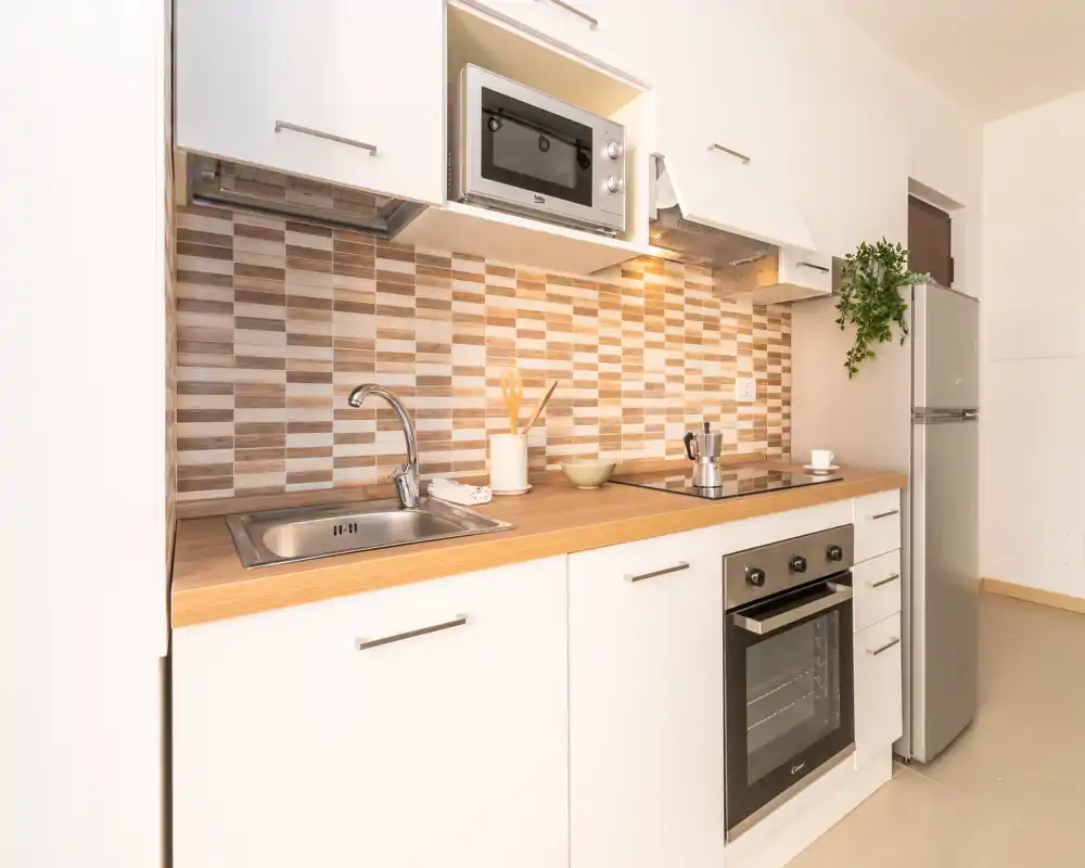 Modern compact kitchen with wood-effect countertop, stainless sink, built-in oven and fridge; warm tiled backsplash and clean white cabinets create a bright, functional space.