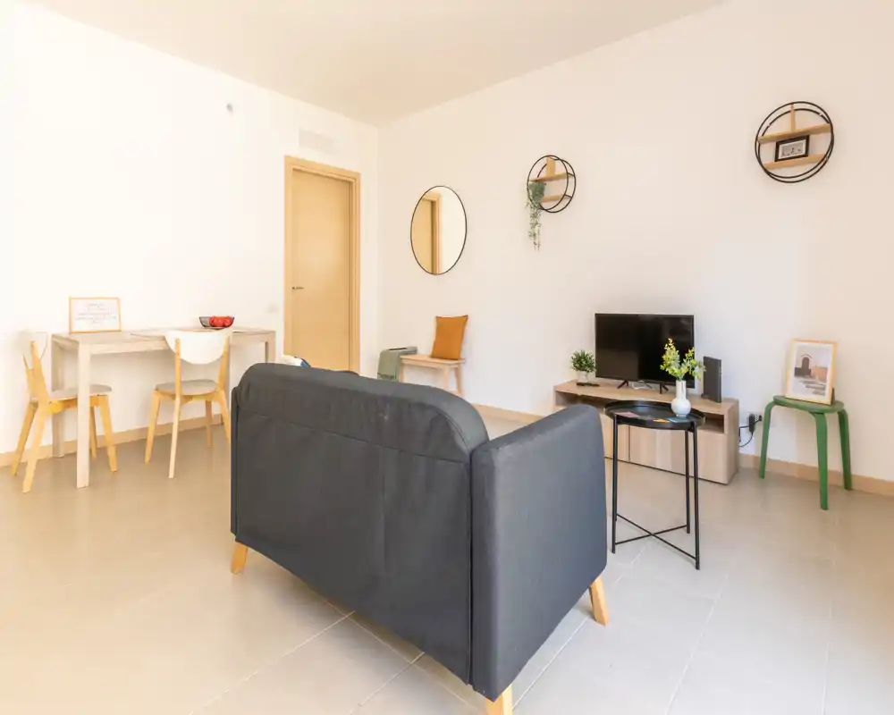 Bright, modern living area with a compact dark sofa facing a small TV unit, minimalist dining nook, and decorative wall shelves — ideal for a cozy apartment stay.