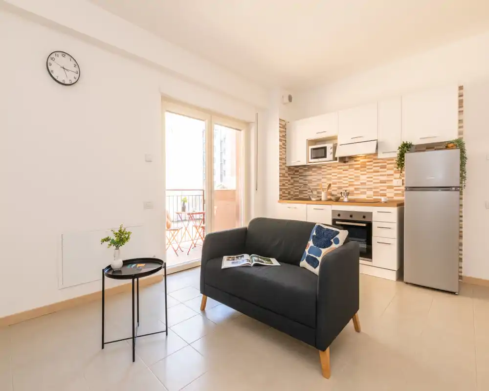 Bright, modern open-plan living area with a compact kitchen, a dark two-seater sofa and a balcony door letting in natural light — ideal for a cozy city stay.