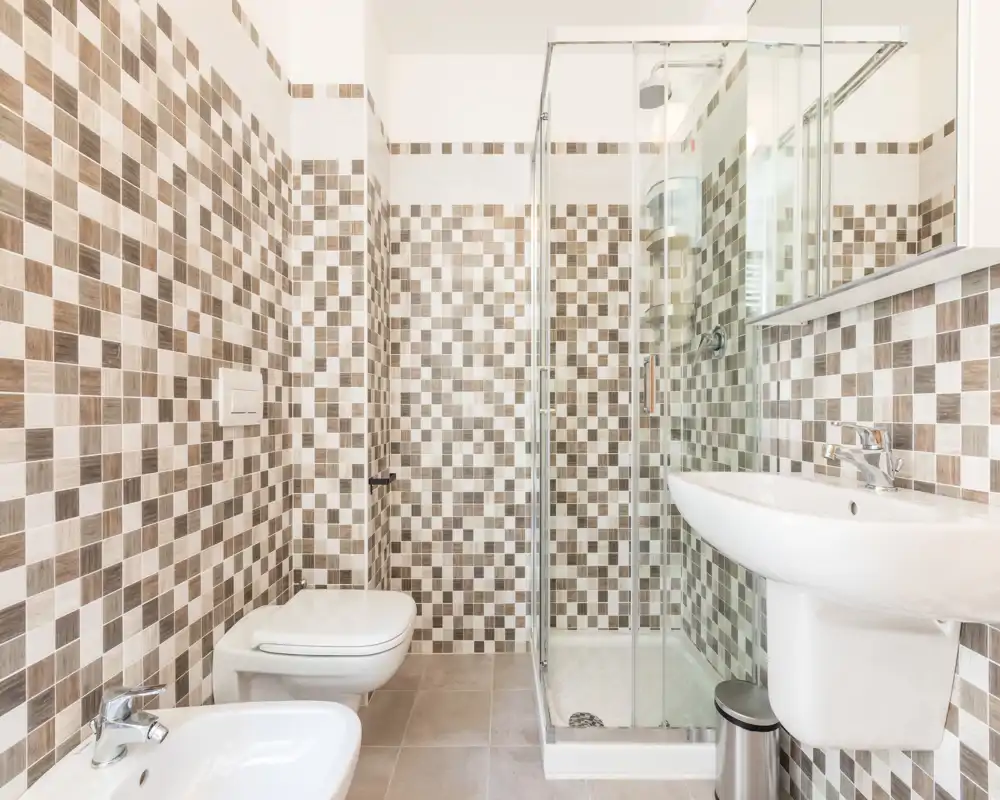 Bright, modern bathroom with mosaic tile walls, a glass-enclosed shower, wall-hung toilet, bidet and a pedestal sink — clean and well-lit.