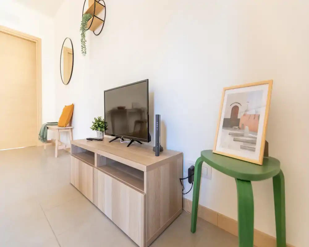 Bright and minimal living area with a low wooden TV console, flat-screen TV, decorative mirrors and a green stool with framed art. Clean tiled floor and light walls create a calm, modern atmosphere.