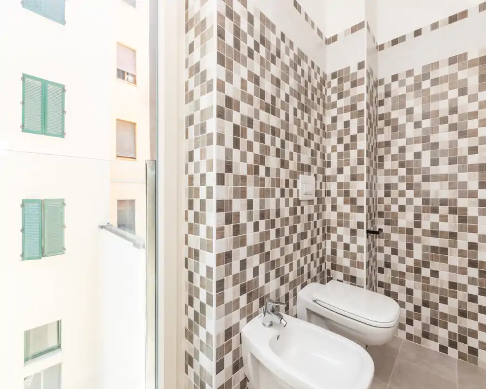 Bright, modern bathroom corner with patterned mosaic wall tiles, a bidet and a wall-mounted toilet under good natural light from a nearby window.