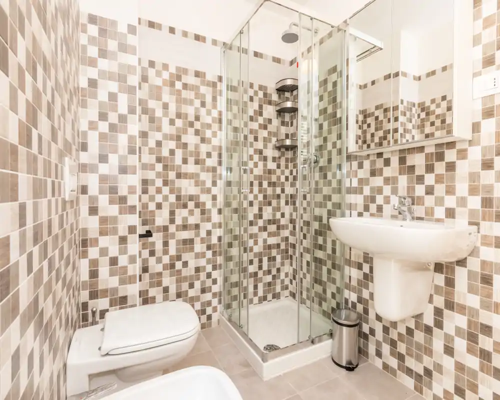 Bright, modern bathroom with a glass-enclosed shower, wall-mounted sink, bidet and toilet, finished in neutral mosaic tiles.