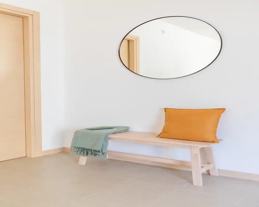 Minimal, well-lit entrance with a wooden bench, decorative cushion and oval wall mirror—clean and inviting hallway corner.