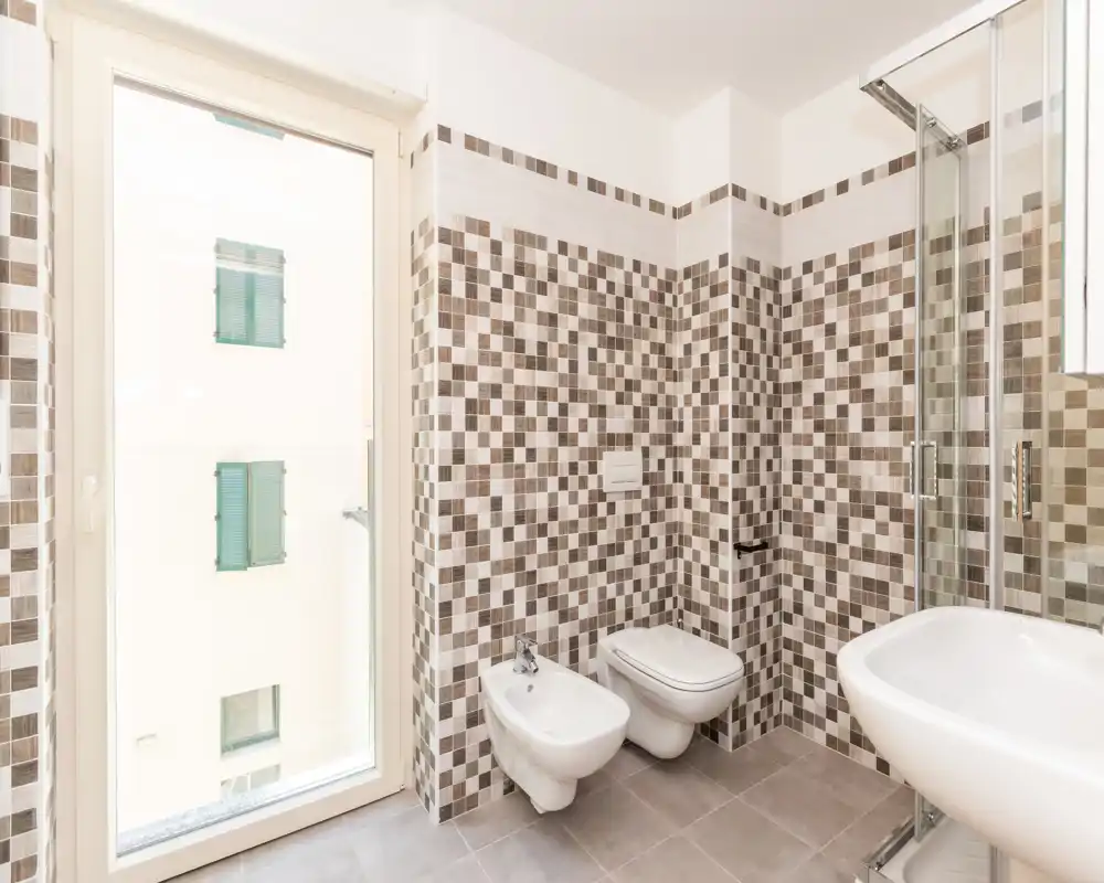 Bright modern bathroom with mosaic wall tiles, wall-mounted toilet and bidet, glass shower enclosure and large window providing natural light.