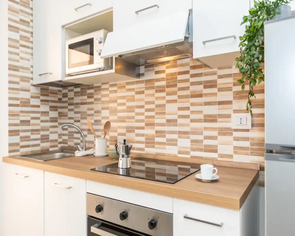 Modern compact kitchen with a wooden countertop, induction hob, built-in oven and stainless-steel sink. Warm neutral tile backsplash and small decorative plant create a tidy, inviting cooking space.