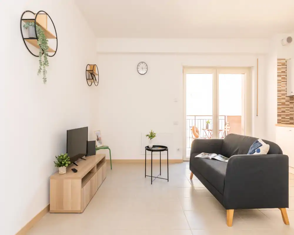 Bright, modern living room with a comfortable dark sofa, compact TV unit and natural light from a balcony — ideal for relaxing or entertaining.