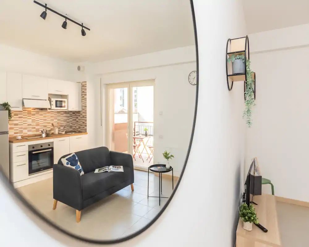 Bright, modern living area with a small sofa facing a compact kitchenette and a balcony. Clean styling, decorative cushions and plants add a welcoming touch — ideal for a city apartment listing.