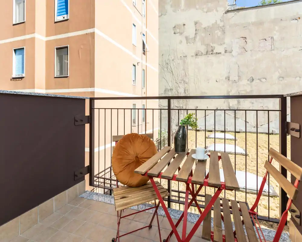 Sunny private balcony with a small wooden bistro table, two folding chairs and a decorative cushion—ideal for morning coffee.