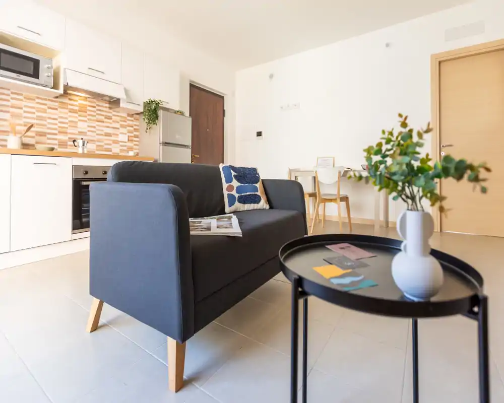 Bright, modern living area with a compact sofa, coffee table and open kitchenette; nicely staged for a rental listing with simple, contemporary decor.
