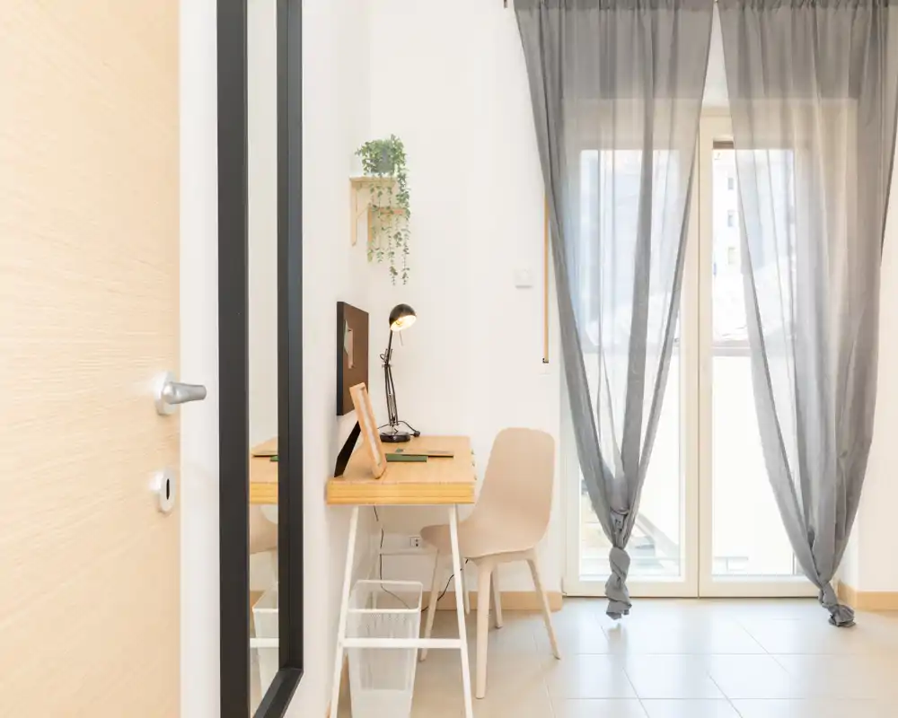 Bright, minimal room with a small desk and chair by a glass door with sheer curtains; suitable as a compact living area or workspace with clean, modern styling.