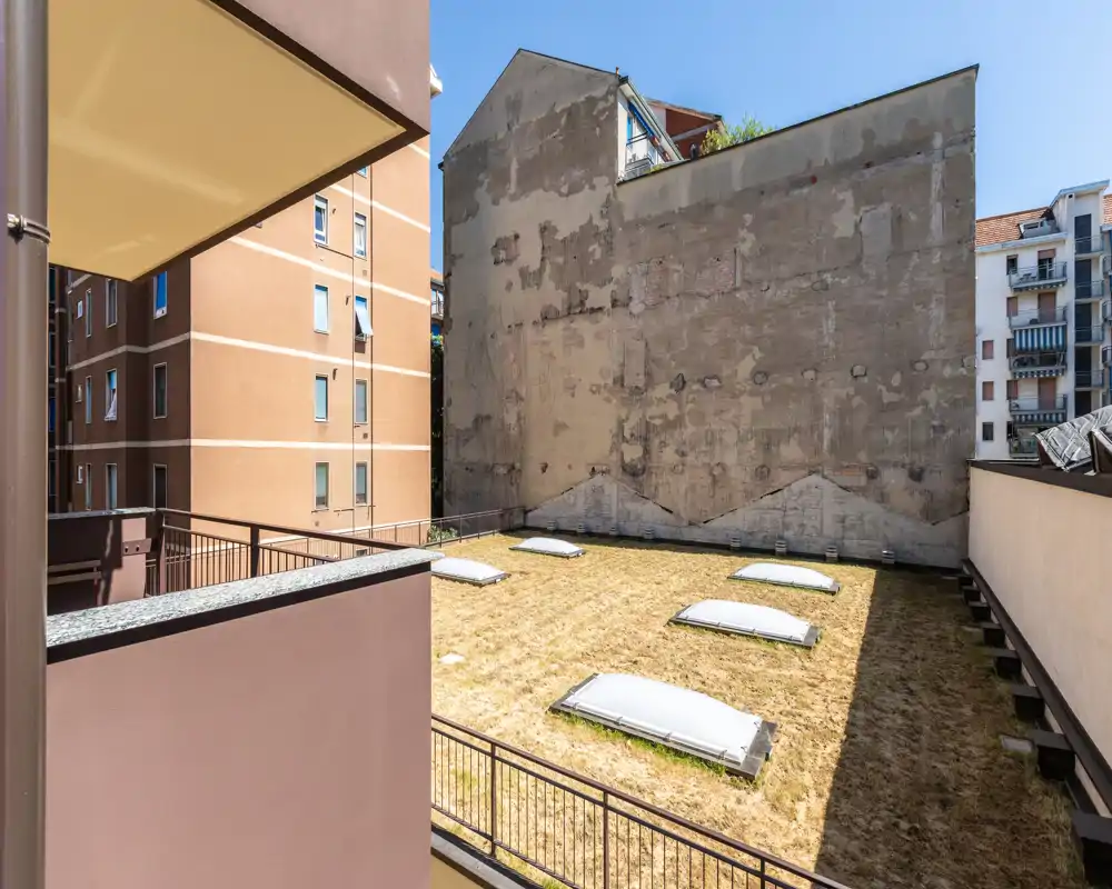Exterior view of a low rooftop/terrace with skylights set in dry grass, surrounded by neighboring apartment walls and railings — bright midday light.
