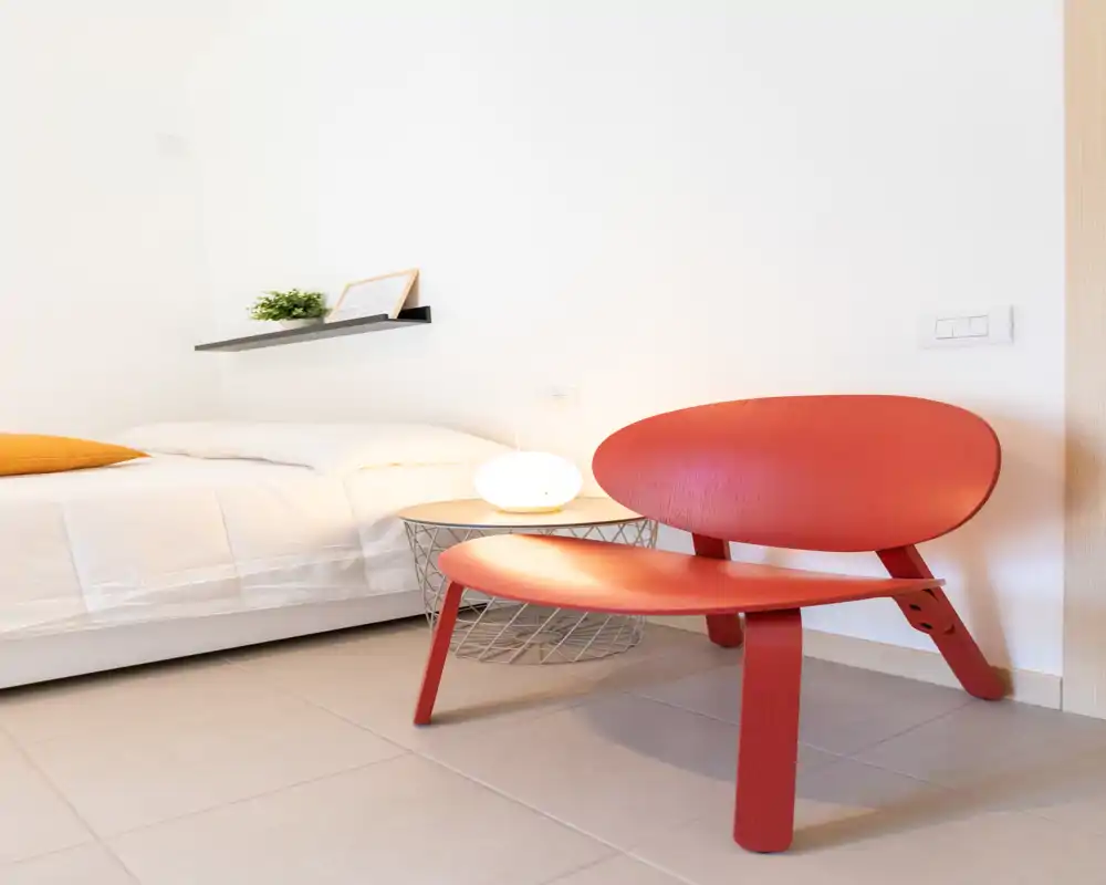 Bright, modern bedroom corner with a white bed, bedside table with lamp and a low red designer chair creating a cozy sitting area.