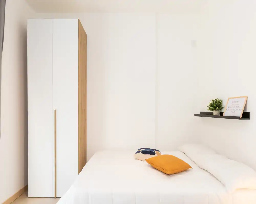 Bright, minimal bedroom with a double bed, soft white linens and a compact wardrobe beside it. Simple decor includes colorful cushions and a small shelf with plant and frame, creating a calm, airy atmosphere.