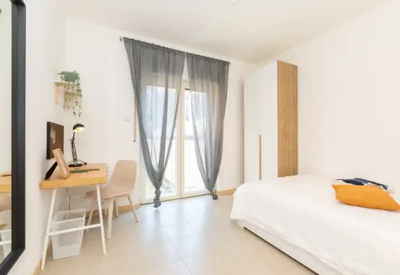 Bright, minimal single bedroom with a neatly made bed, a wooden wardrobe and a small desk by the window letting in abundant natural light.