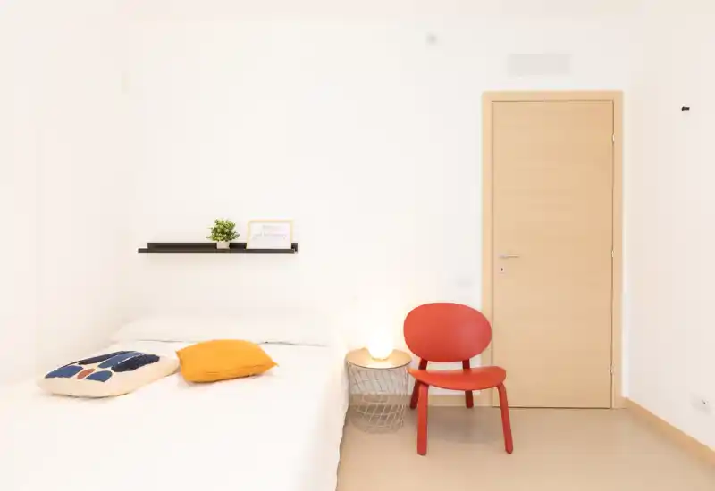 Compact, bright bedroom with a double bed, accent pillows, a small bedside table with lamp and a red chair — minimal, modern styling and lots of natural-white light.