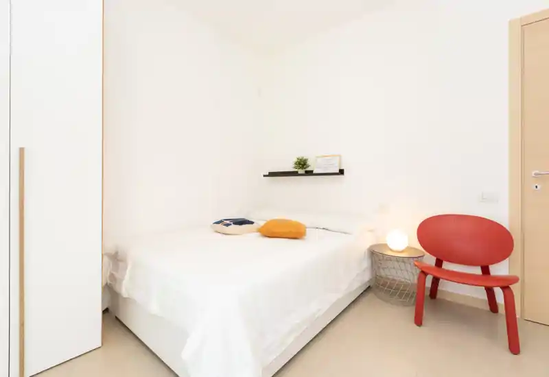 Bright, minimalist bedroom with a double bed dressed in white linens, decorative cushions, a small wall shelf and a red accent chair — ideal for a modern short-term rental.