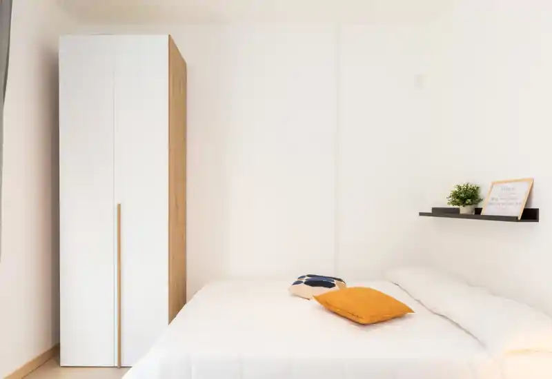 Bright, minimal bedroom with a double bed, soft white linens and a compact wardrobe beside it. Simple decor includes colorful cushions and a small shelf with plant and frame, creating a calm, airy atmosphere.