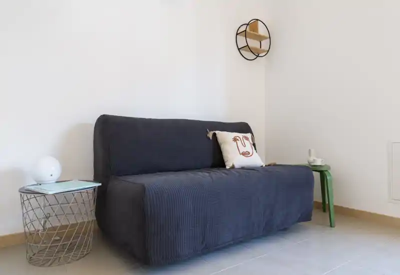 Compact living area with a dark upholstered two-seater sofa, decorative cushion and two small side tables offering a minimalist, cozy vibe and clean, neutral walls.