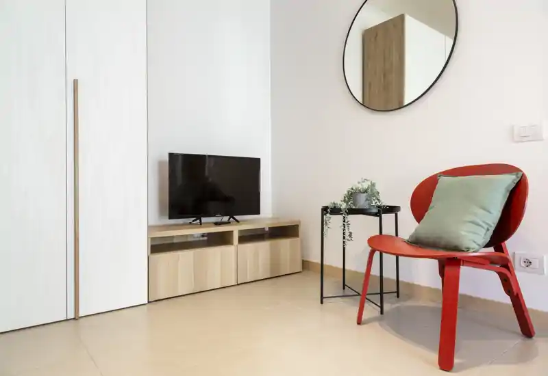 Minimalist living area with a low wooden TV console, a red accent chair with a green cushion, a small side table with plant, and a round wall mirror — bright, tidy and modern.