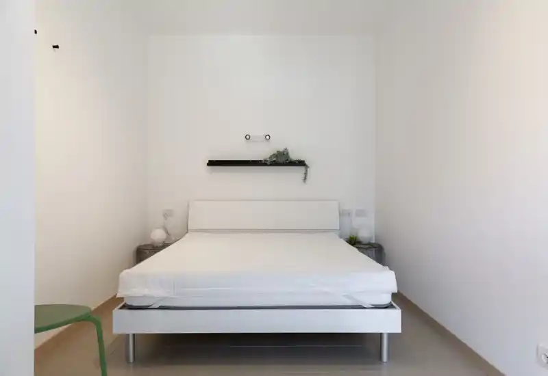 Minimalist bedroom with a white platform bed centered against a plain wall, two small bedside tables and subtle decorative greenery for a calm, bright atmosphere.