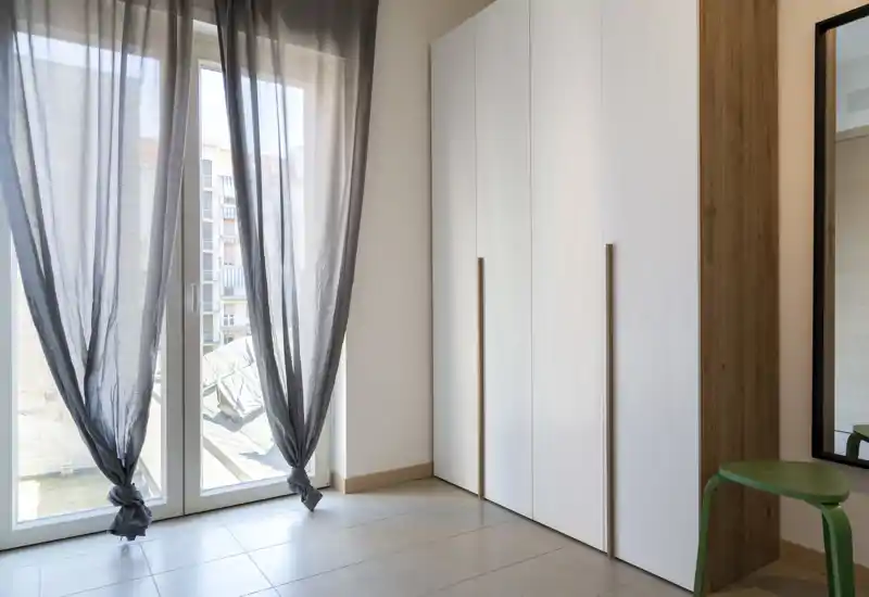 Bright corner with large glass doors, sheer curtains and a tall white wardrobe; simple, modern finishes and tiled floor create a clean, airy space.