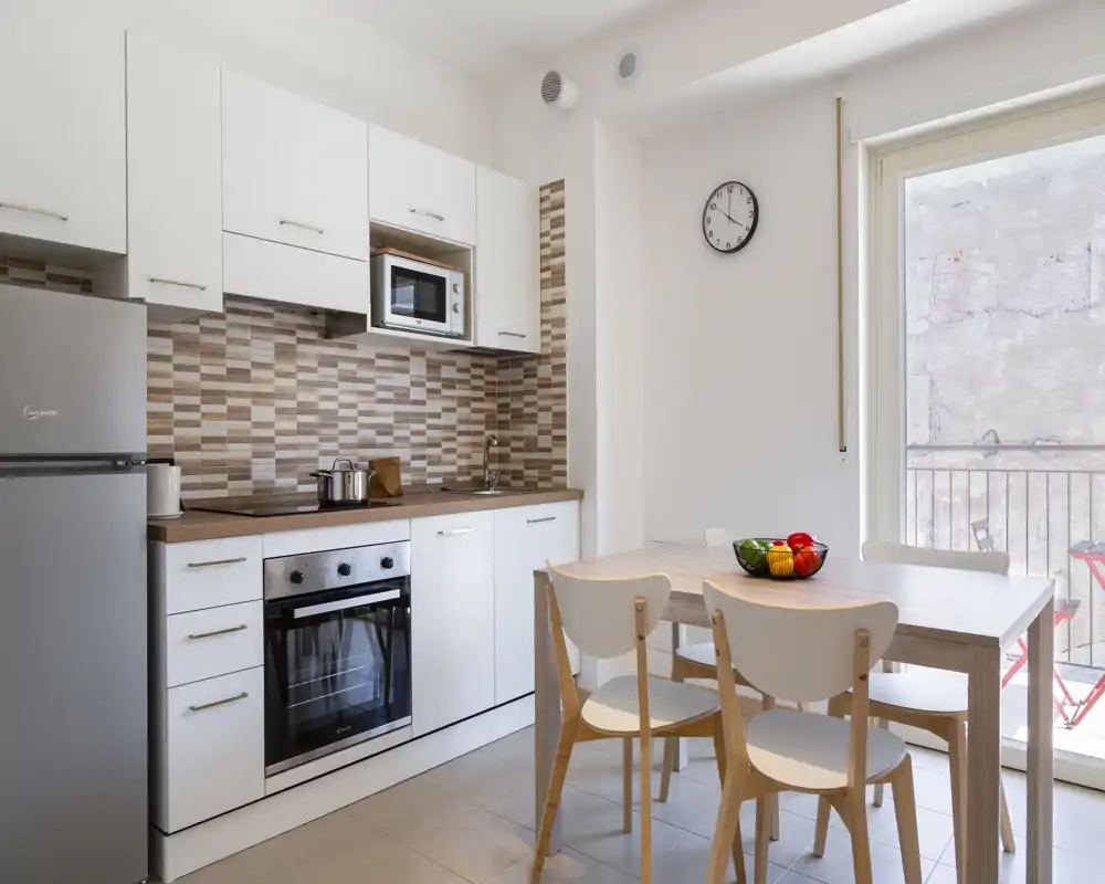 Clean, modern kitchenette with white cabinets, built-in oven and microwave, a wooden dining table with four chairs, and a bright balcony door letting in natural light.