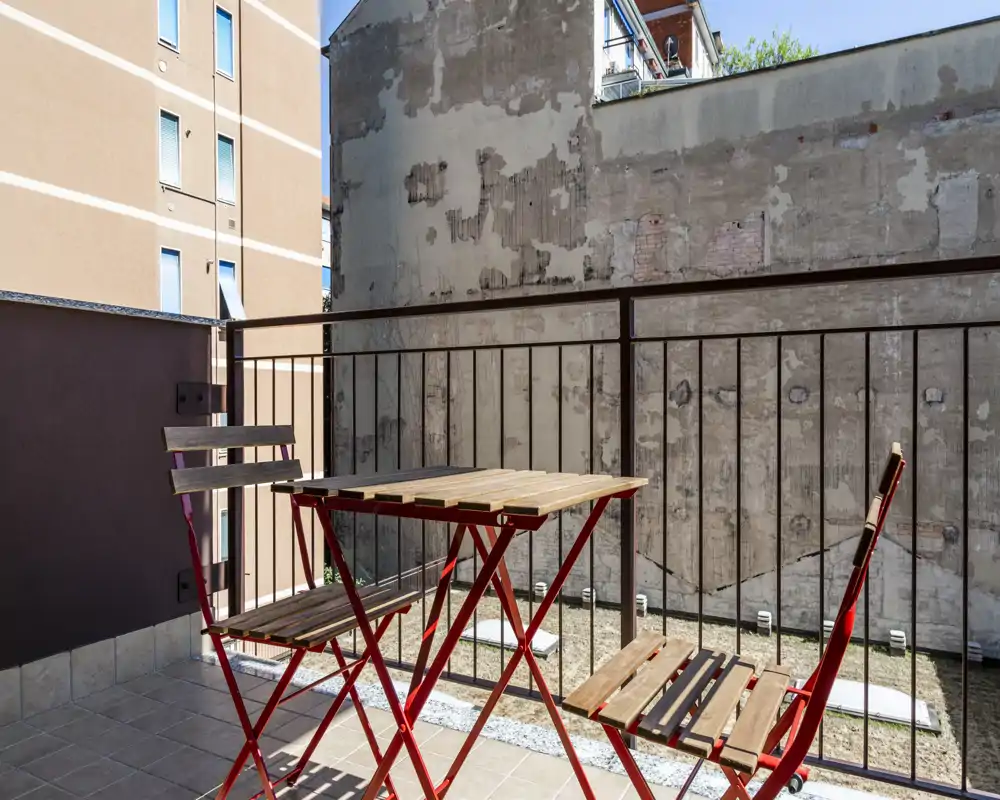 Small sunny balcony with a folding wooden bistro table and two matching chairs; tiled floor and metal railing overlooking neighboring buildings.