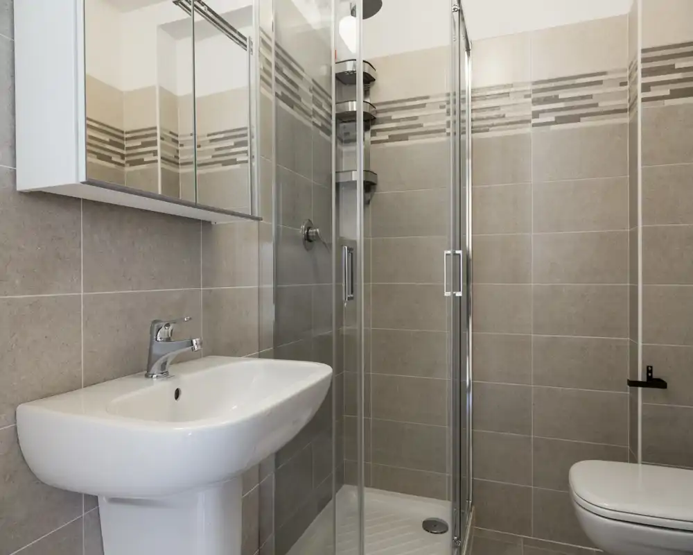 Modern, compact bathroom with a glass shower enclosure, pedestal sink and wall-mounted toilet; neutral beige tiles and a mirrored cabinet complete the clean, contemporary look.